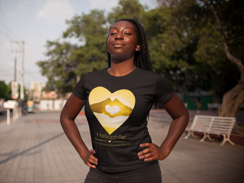 a woman of color with her hands on her hips looking up with the warmth of the sun on her face, wearing a t-shirt saying 'heliophile - a person who loves the sun' and a pair of hands making a heart against a bright yellow sun