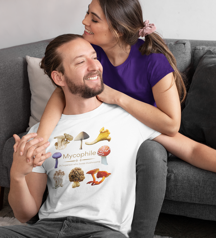 A man leaning against a woman on a couch, both smiling, he is wearing a t-shirt with 8 different types of real mushrooms such as morel and chanterelle; it also says 'mycophile - a person who loves mushrooms'