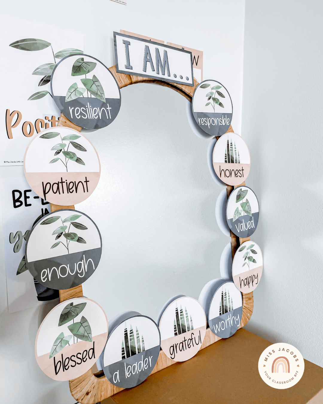 On the left, we see a series of white magazine holders, each with a circular label on them. The label features children’s names and a plant illustration. On the right we see the display created in an office setting - an arch-shaped mirror is surrounded by circular affirmations each with a plant illustration. The top of the mirror says ‘I am and the affirmations include enough, patient, a leader, grateful and happy.