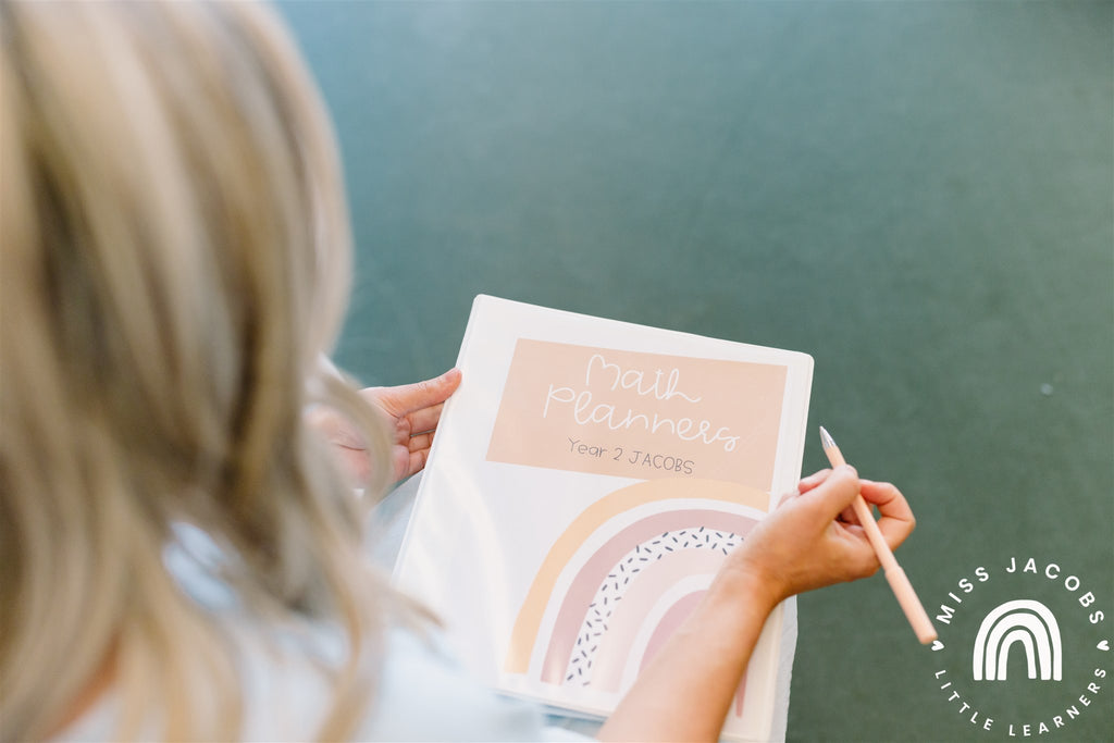 The image is taken over the shoulder of Chantelle, who sits with a white binder on her lap. The cover features a rainbow graphic and the words ‘Math Planners, Year 2 Jacobs.’ She holds a pale peach coloured pen in her hand.