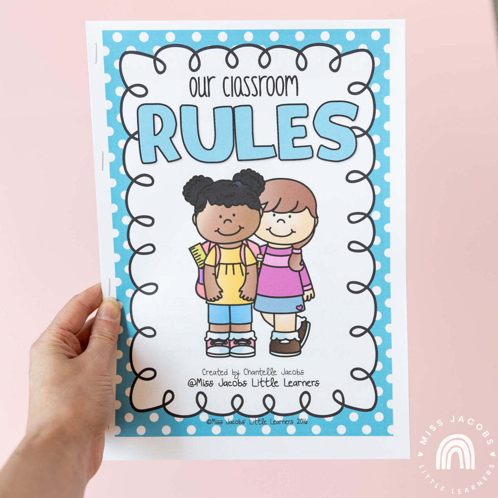 A hand holds an illustrated student booklet that reads ‘Our classroom rules,’ above two illustrated school children. The booklet has a blue border with white spots.