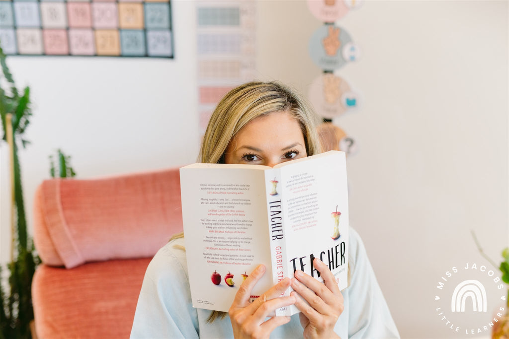 Chantelle sits on a peach coloured chair in a bright white classroom, she peeks over the top of a book titled ‘Teacher.’