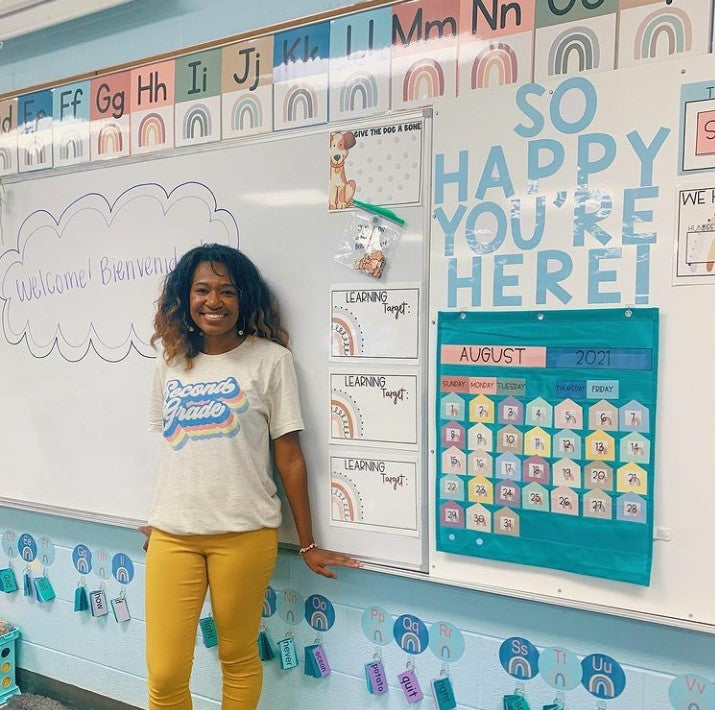 Educator Hailey stands in front of a whiteboard featuring a range of different displays in shades of blue and teal. The Boho Rainbow Alphabet Posters line the top of the board. Blue letters spell ‘So happy you’re here!’