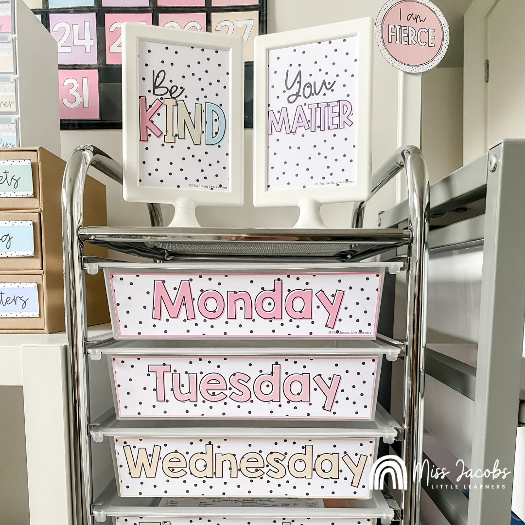 Two images are side by side, on the left we see a teacher's toolbox, which is a small cube with nine little plastic drawers. Each of the drawers is labelled, using items from the Spotty Pastels range. The labels are in pink, yellow, green and blue with small black dots on a white base. The drawer’s labels include notes, pins, erasers and clips. on the right we see a teacher’s trolley, a 10 drawer metal trolley, with spotty pastel labels on the front of each draw, listing the days of the week. On top of the trolley sits two framed posters, one saying ‘be kind’ and the other saying ‘you matter’. Both in Spotty Pastels colour palette and style