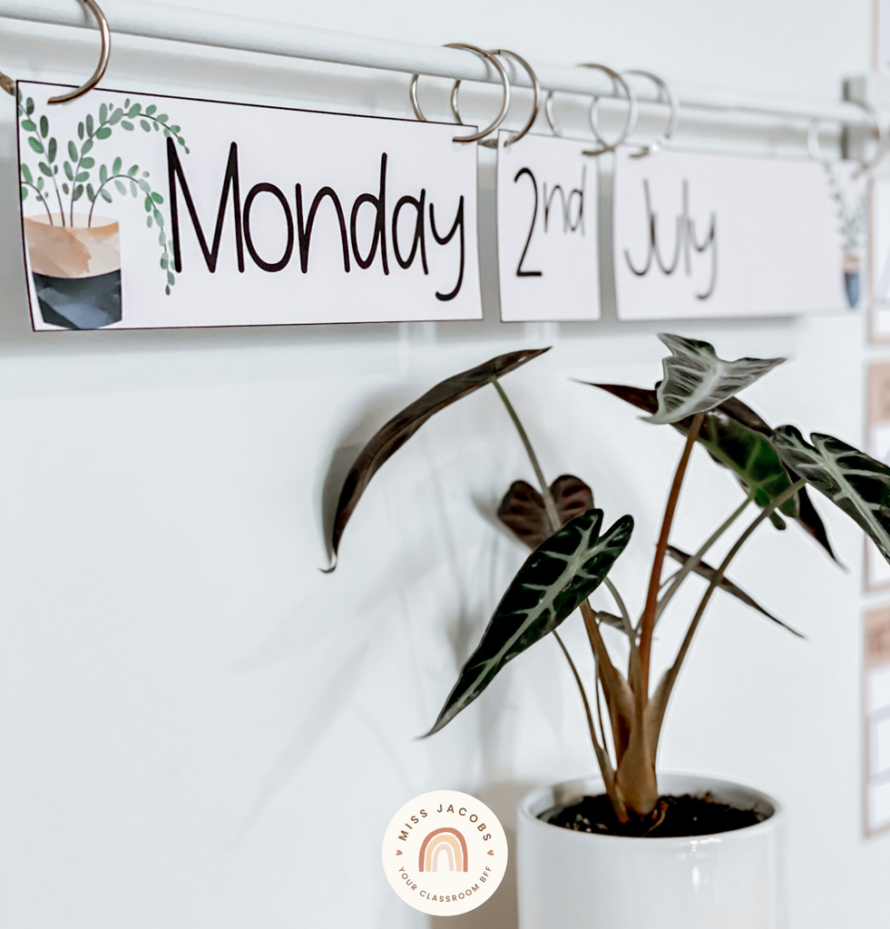 A rod is mounted on a whiteboard to create a classroom flip calendar. Three words hang from the rod - the name of the day, the date and the month. They’re affixed to the rod with large rings and can be flipped over.