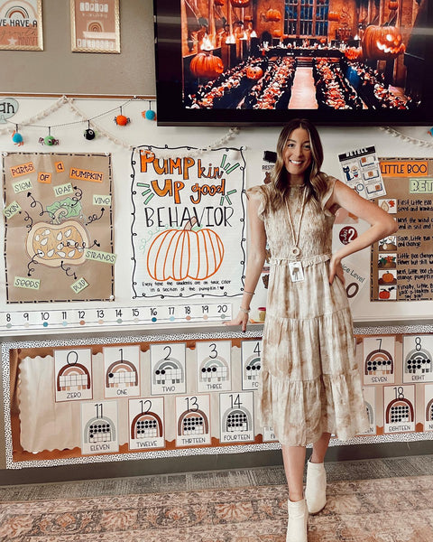 A teacher stands in front of a whiteboard filled with display items in a neutral palette with orange highlights. We see a diagram of a pumpkin, and a poster with the words ‘PUMPkin up good behaviour.’ There’s also a TV screen with the Halloween scene from Harry Potter.