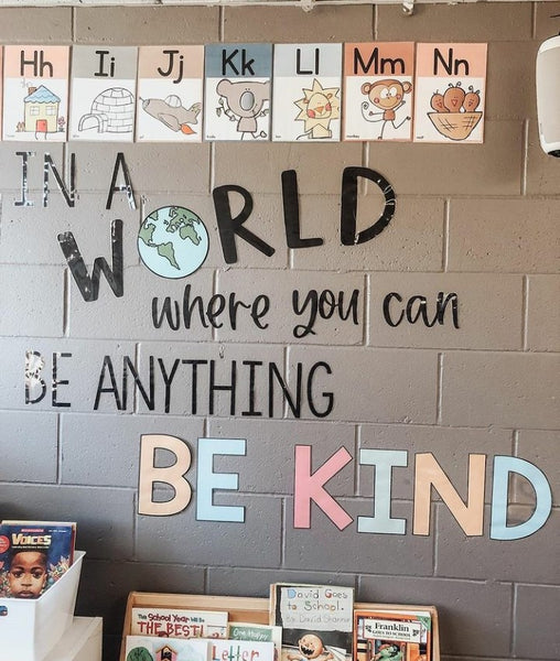 A grey brick wall features cutout letters that read ‘In a world where you can be anything, be kind.” At the bottom of the shot you can see the top of a little bookshelf.