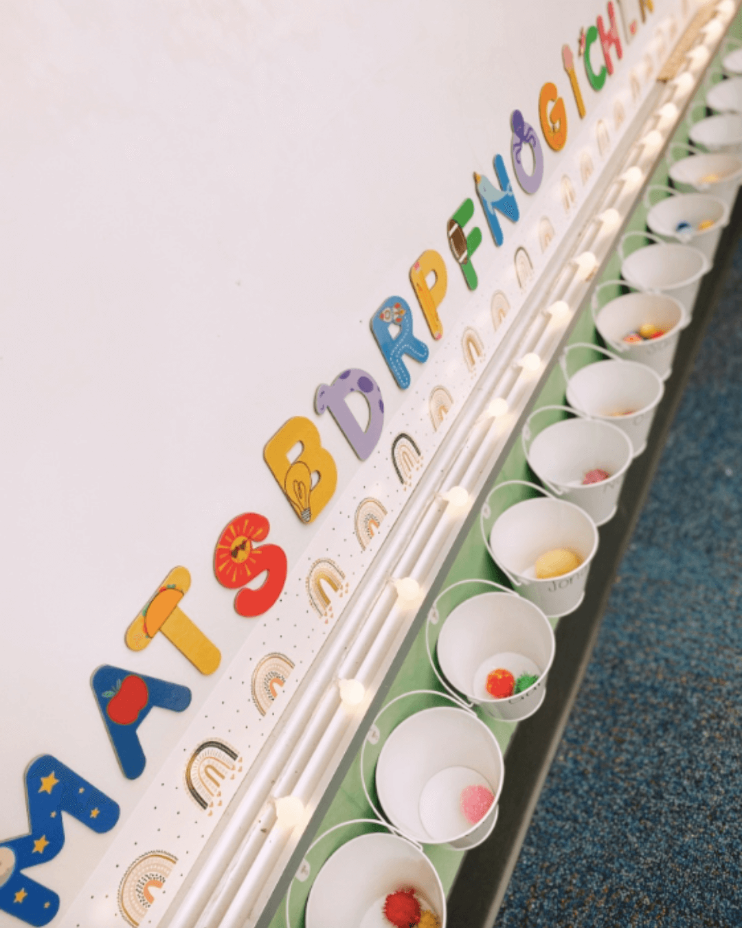 The left image shows Sabrinna holding a bucket in front of her whiteboard with decor from the Modern Rainbow range. The right image shows the small white buckets hanging underneath the whiteboard, with pom-poms inside.