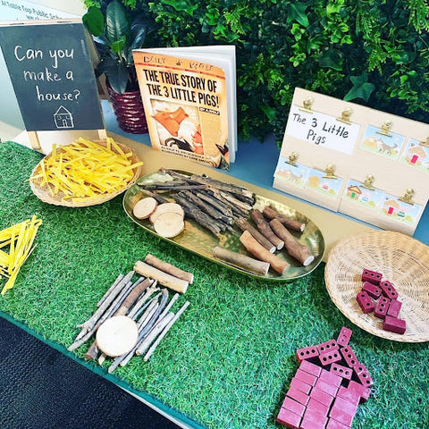Two tables are covered with artificial turf. On top of them are a range of found materials and objects that bring to life the relevant books - with a sea life theme on the left, and a Three Little Pigs theme on the right.