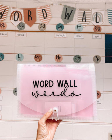 Alt text: In the background of the image is a light-coloured brick wall. Along the top of the wall, colourful bunting is strung that reads ‘Word Wall,’ with circular labels underneath that have the letters of the alphabet printed on them. In the foreground of the image, a hand holds up a pink folder that reads ‘word wall words.’