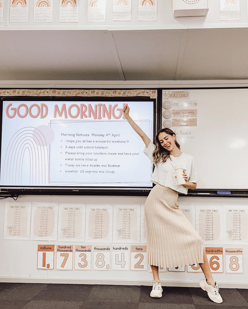 A teacher stands in front of the digital whiteboard with her arm in the air in celebration. The classroom is a mixture of earthy tones.