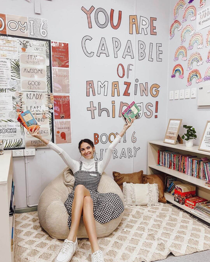 A classroom corner features a textured rug in neutral tones, with a cream coloured bean bag and @miss_grosse_ sitting on it holding books up. She is surrounded by a cute bookshelf and a wall display that reads ‘you are capable of amazing things’ in Miss Jacobs Boho Vibes Neutral letting pack.