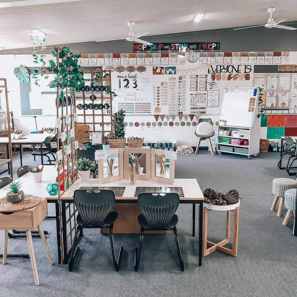 A wide shot of a classroom shows desks, whiteboards and a variety of chairs. In the foreground, a science and nature zone includes a lattice with creeping ivy, a desk with mirrors and a side table with pinecones. The whiteboard and back wall features items from the Boho Vibes Decor Bundle in Desert Neutral Tones.