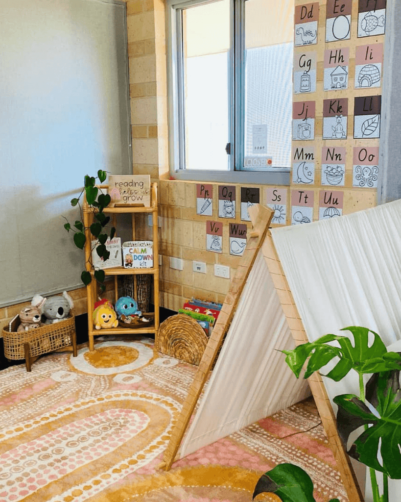 A classroom corner features a small white fabric tent, and a rug in neutral tones, with an Indigenous art print. The wall features alphabet posters from my Spotty Neutral range. There are a number of plants and a small cande bookshelf. All of the tones match, including the sandy coloured exposed brick wall.