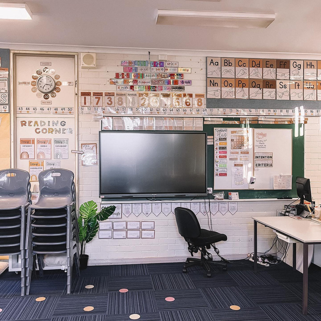 The image shows a classroom wall with a large screen and a number of MJLL wall displays from the Boho Neutrals range. We can see a clock, alphabet and number posters, a desk and a stack of grey chairs.