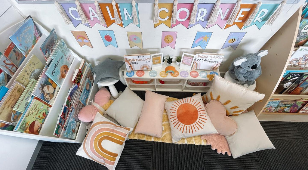 A birds’ eye view of a reading corner in a classroom shows bookshelves and a variety of cushions in neutrals and pinks. The words ‘Calm Corner’ are created through pastel coloured bunting letters. There are small signs of affirmations on a small shelving unit.