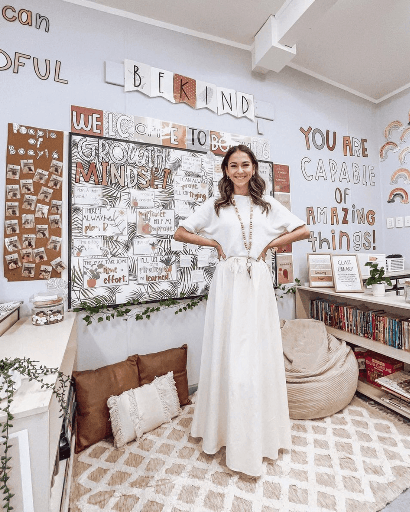 A teacher wears a white top and skirt and stands in front of a beautiful classroom. Neutral tones and displays create a warm and inviting atmosphere. We see the words ‘you are capable of amazing things’ and other affirmations spelled out on the walls.