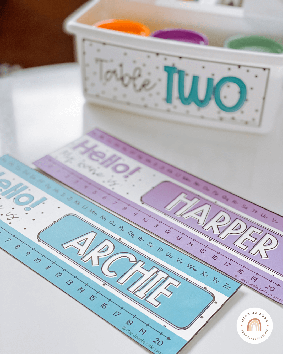 Two images show name tags on a desk, and on a series of white tubs on a trolley. The labels are in purple, blue and green with a spotted background.” style=