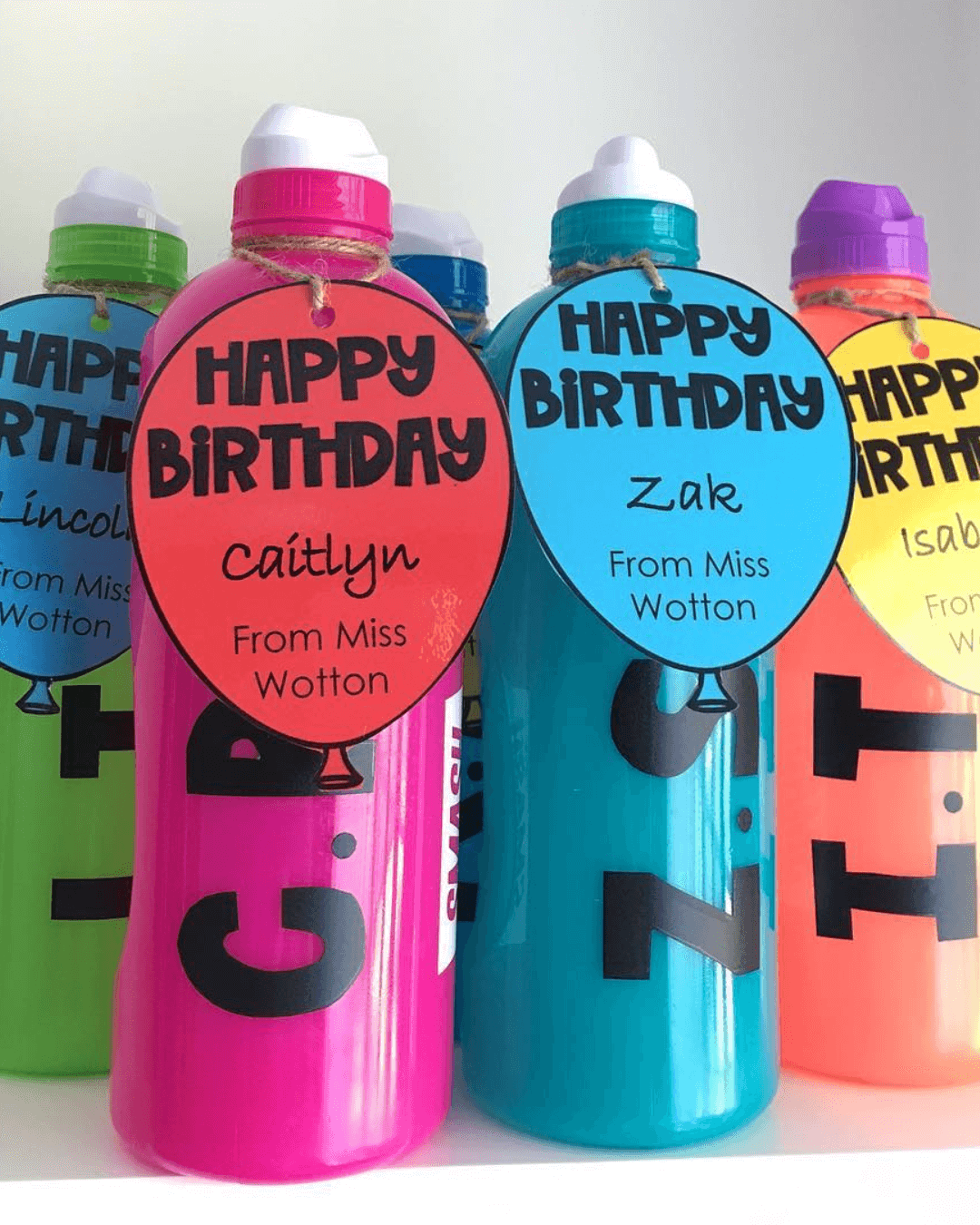 The left image shows the happy birthday balloon labels,  affixed to customised colourful drink bottles. The right-hand image shows the same balloons tied to small plushie toys including a leopard, a duck and a pig.” style=