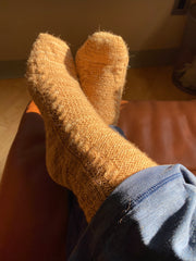 A persons feet crossed over one another and propped up on a stool, they’re wearing handspun handknit socks with cables up the sides in a burnt orange color.