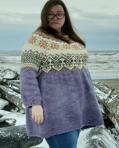 Madeline, a fat pale woman with long brown hair and glasses, stands on a wind swept snowy beach with waves in the background.  She wears a color work yoke sweater dress made with naturally dyed yarns.
