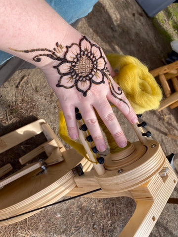 A photo of Madeline’s hand with a henna tattoo of a sunflower done by Julian on it, she holds a bit of naturally dyed wool she is working on spinning and her wheel can be seen in the background.