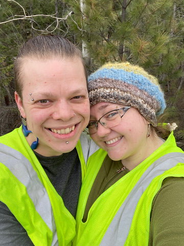 Julian (left - a pale trans man with his hair pulled back) & Madeline (right a fat pale woman wearing a handspun rainbow hat and glasses) lean into eachother smiling while they wear matching safety vests and stand in front of a pine forest background.