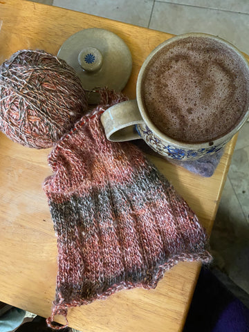 Madelines knitting, a simple scarf in an autum pallet in hand spun yarn, sits next to a full mug of coffee with a heart in the foam on top.