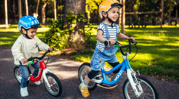 Zwei glückliche kleine Jungen fahren leichte Laufräder: einer hat ein rotes Laufrad Woom 1 und der andere ein blaues Woom 1 Plus