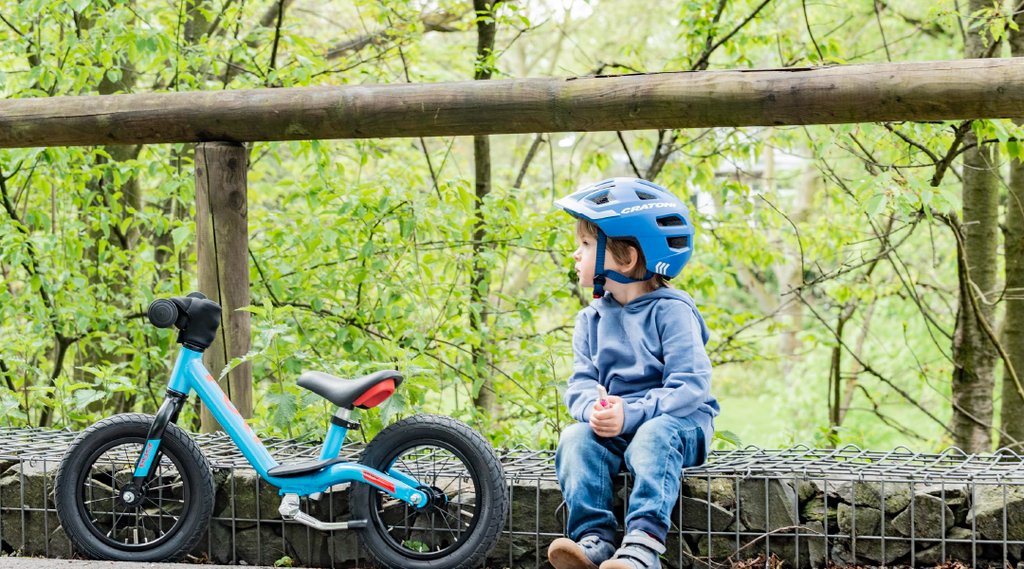 Kleiner Junge sitzt neben seinem blauen Laufrad im Wald