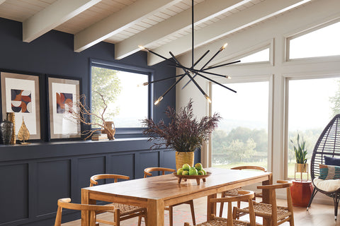 A six light black chandelier over a light wooden table with dark blue walls.