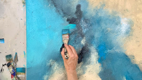 Türkis Blau und Sand auf Leinwand. Abstraktes Kunstwerk an dem eine Hand einen Pinsel hält