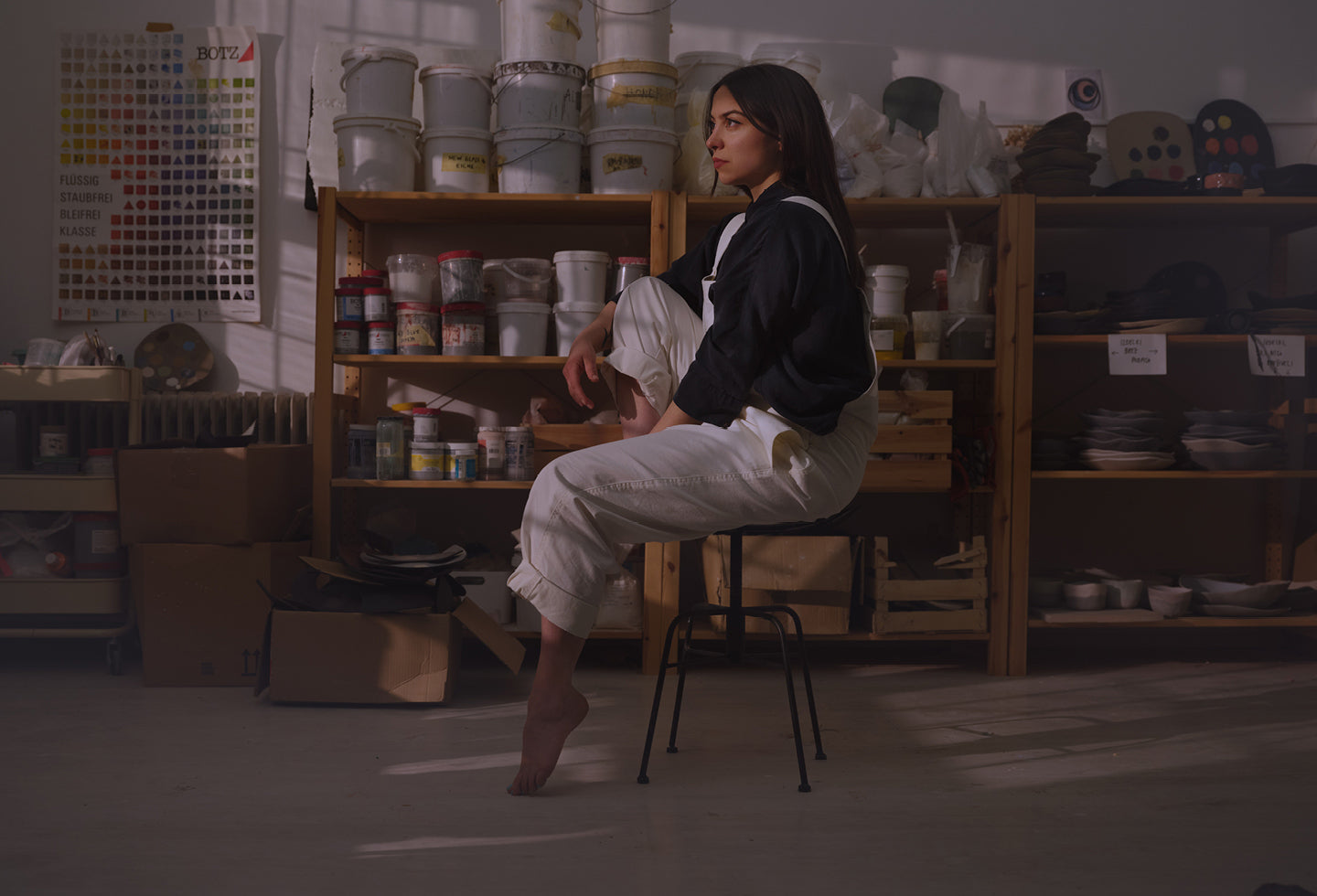 Portrait of Hana Karim in her studio seated on a chair and facing to the left, with shelves of glazes seen in the background