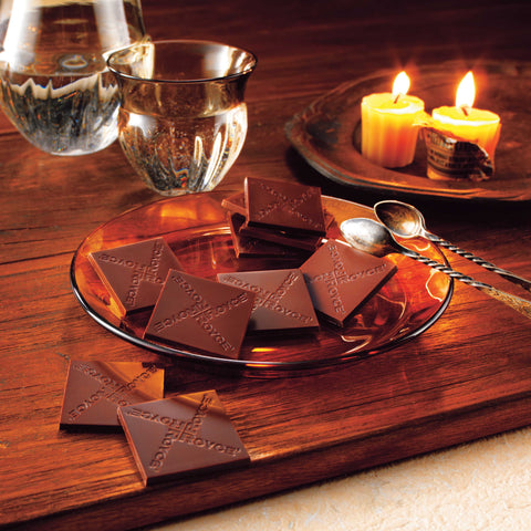 Image shows dark chocolate squares on a plate with wooden board and candles.