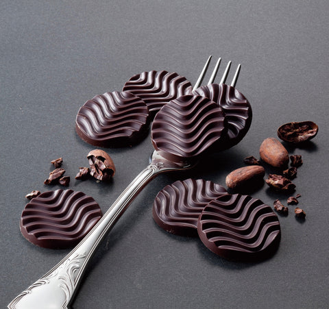 Image shows dark chocolate discs with loose cacao beans and a silver fork.