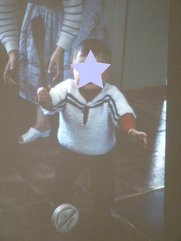 why I knit. Baby standing with arms wide wearing a hand knitted garter stitch top in white with dark blue stripes