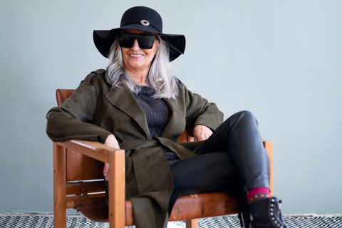woman smiling with silver gray hair wearing a large brim black hat, military green jacket, gray pants and black combat boots sitting in chair