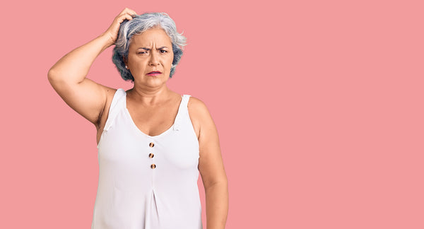 woman with short gray hair scratching her scalp with a confused expression on her face, wear a white sleeveless blouse, pastel pink background