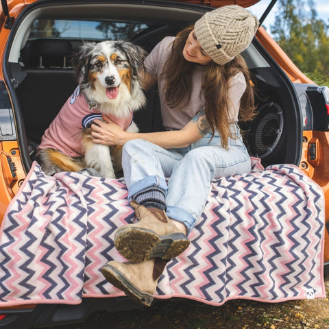 A lady and her dog sitting on a blanket in the back of the car