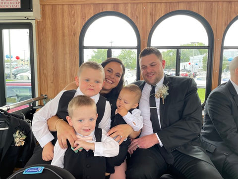 Katie Tufts, Chief Operating Officer & General Counsel of The Bold Bow Tie, sits on a trolley with her husband and three sons who wear their blush bow ties with their ring bearer suits for a wedding. 