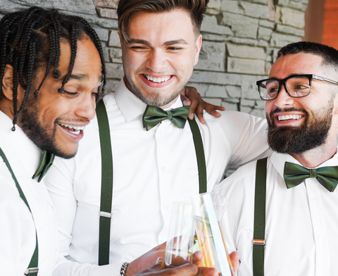 three groomsmen wear satin olive bow ties and olive suspenders and toast with champagne