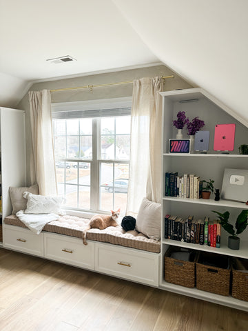 The home office of KDigitalStudio after renovations. There is a large window, which is framed by a window seat and bookshelves. A cat seats on the window seat and looks out the window.