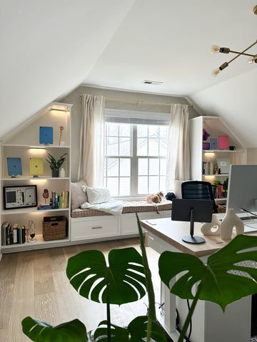 The home office of KDigitalStudio after renovations. There are large windows, which are framed by a window seat and symmetrical bookshelves on either side. In the middle of the room is a desk with an Apple Studio display monitor. Closest to the frame are big, bright green monstera leaves.