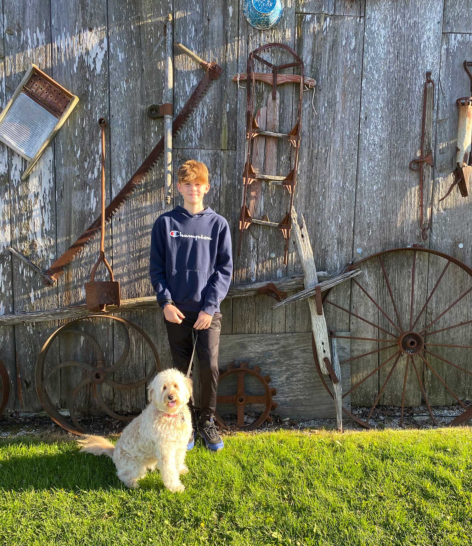young red headed boy in front of old wooden building with antique wheels and tools hanging on walls holding mini goldendoodle
