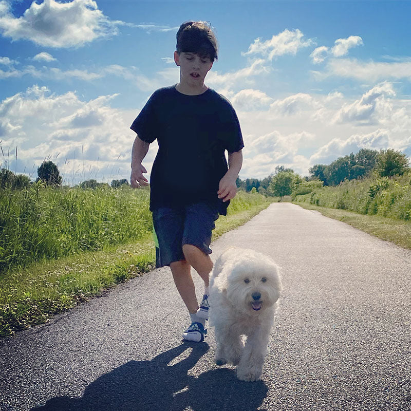 young boy running along side a mini goldendoodle dog on a long paved path with prairie grass on the sides and trees in the background and blue sky with white clouds