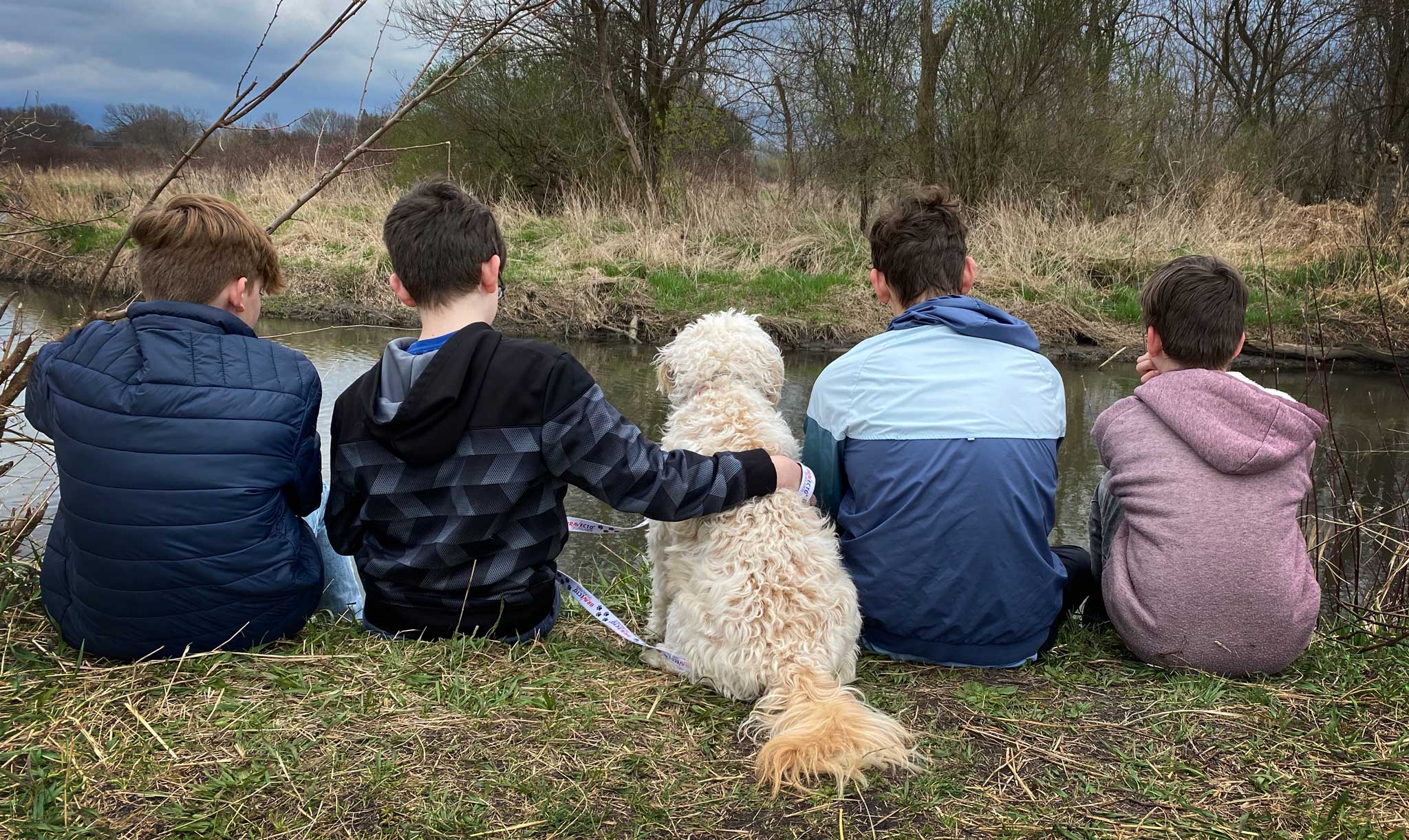 4 young boys in light jackets and hoodies with their mini goldendoodle dog sitting facing a creek