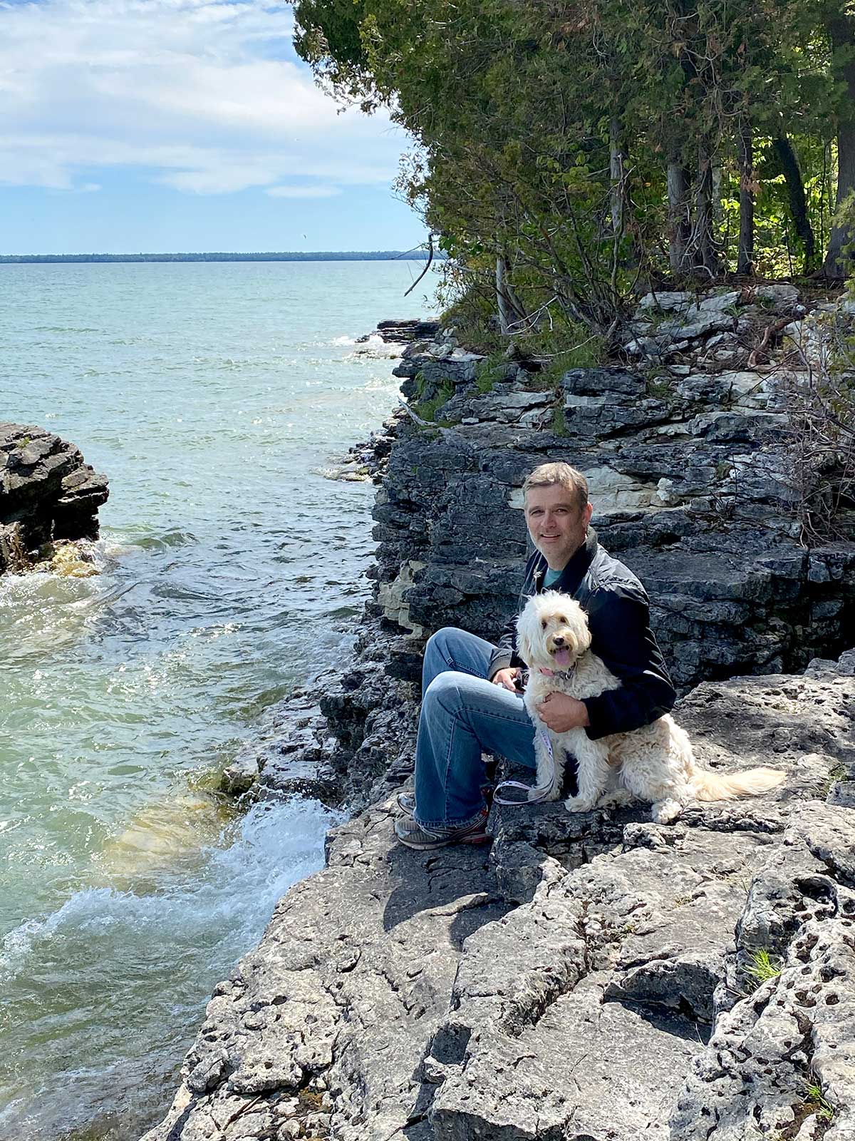attractive man sitting on rocky bluff next to blond mini goldendoodle by Lake Michigan in Door County, Wisconsin