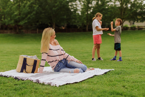 woman sitting on picnic blanket with kids playing in the background