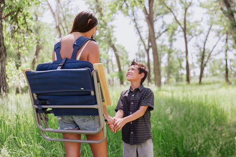 woman carrying chair wit backpack straps while holding her sons hand and walking through a field