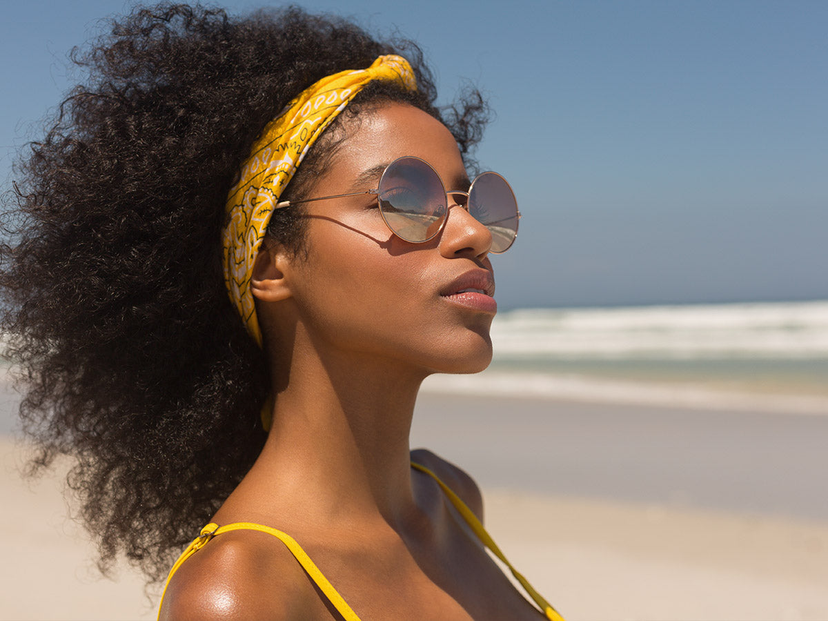  A woman with a large afro held back with a yellow bandana standing on the beach looking away with round sunglasses.